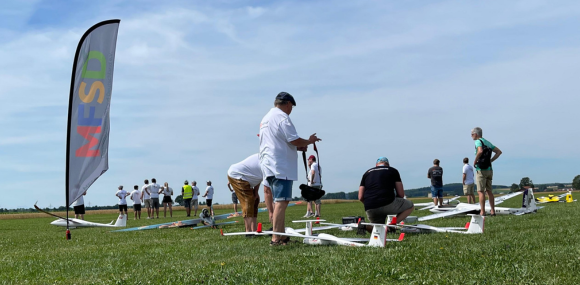 Menschen versammeln sich auf einer Wiese mit mehreren Modellflugzeugen am Boden. Einige bereiten die Flugzeuge vor, andere beobachten das Geschehen. Links im Bild steht eine bunte Flagge mit der Aufschrift "MFSD". Der Himmel ist leicht bewölkt und es herrscht sonniges Wetter.